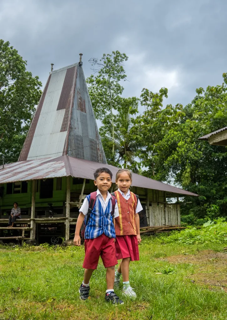 Dua anak tersenyum cerah di depan rumah tradisional, melangkah menuju sekolah dengan penuh semangat—cerminan harapan dan perjuangan anak-anak NTT dalam meraih akses pendidikan yang lebih baik. Foto: © Tim Komunikasi INOVASI 2025